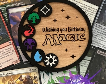 May include: A wooden coaster with a black border and a light brown background. The coaster has a black and white image of a Magic: The Gathering mana symbol in the center. The text "Wishing you Birthday Magic" is written in a black font on the coaster. The coaster is surrounded by Magic: The Gathering cards.