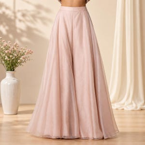 May include: A floor-length, high-waisted skirt in a soft, pale pink hue. The garment features a structured waistband and a voluminous, flowing silhouette crafted from a sheer, layered fabric that creates a subtle, semi-transparent effect.