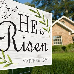 K&ouml;nnte beinhalten: Ein wei&szlig;es Holzschild mit gr&uuml;nen Bl&auml;ttern und einer Taube mit einem Olivenzweig. Das Schild tr&auml;gt die Aufschrift "HE is Risen" mit dem Bibelvers "MATTHEW 28:6".