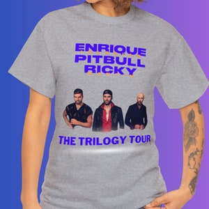 May include: Gray t-shirt with a purple print of Enrique Iglesias, Pitbull, and Ricky Martin. The text "The Trilogy Tour" is printed below the images.