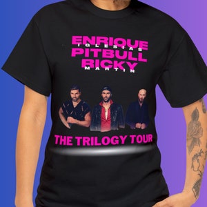 May include: Black t-shirt with a pink graphic of Enrique Iglesias, Pitbull, and Ricky Martin. The text "The Trilogy Tour" is below the image of the singers.