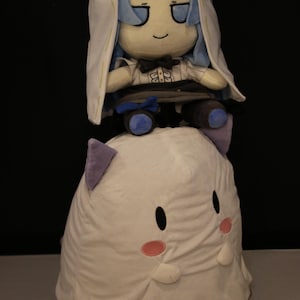 May include: Two plush toys, one stacked on top of the other. The top plush is a doll with blue hair and a white hood. The bottom plush is white with purple ears and pink cheeks. The toys are on a dark background.