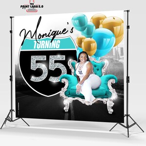 May include: A birthday backdrop featuring a woman sitting on a turquoise and silver throne. The backdrop has a black and turquoise highway sign with the text "Monique's Turning 55" and a background of a city skyline. The backdrop is decorated with blue and gold balloons.