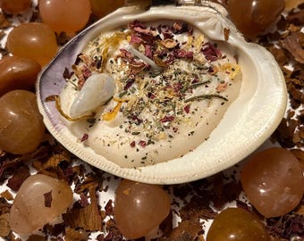 May include: A white seashell candle with dried flowers, herbs, and a clear crystal inside. The candle is surrounded by small orange-brown stones and wood chips.
