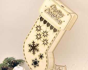 May include: A wooden Christmas stocking-shaped box with laser-cut snowflake designs and the words "Merry Christmas." The box is made of light-colored wood and has a small, separate wooden box next to it. The background includes white candles and pinecones.
