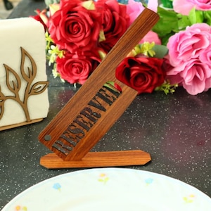 Op de afbeelding: Een houten bord met het woord "GERESERVEERD" in zwarte letters. Het bord staat op een tafel met een witte servet houder en een boeket rode en roze rozen op de achtergrond.