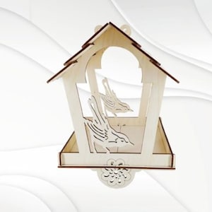 Puede incluir: Un comedero para pájaros de madera con forma de casa y un diseño de pájaro decorativo. El comedero tiene una pequeña plataforma para que los pájaros se posen y un techo para protegerlos de los elementos.