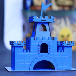 Peut inclure: Un modèle de château en bois bleu avec un drapeau sur le dessus. Le château a une tour, des murs et une porte.