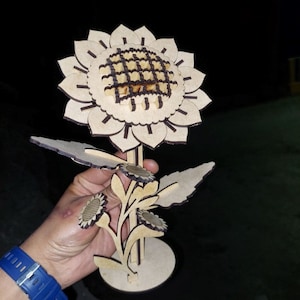 May include: A wooden sunflower sculpture with a brown and beige color scheme. The sunflower has a detailed center and multiple petals. The sculpture is on a stand with three smaller sunflowers.