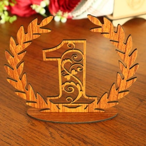 May include: Wooden table number with the number 1 in a decorative font surrounded by a laurel wreath. The number is brown with a wood grain texture.
