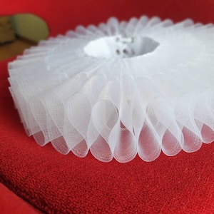 May include: A white, ruffled collar made of sheer fabric. The collar is circular with multiple layers of delicate, wavy ruffles. The item is set against a red background, highlighting the intricate design and texture of the collar.