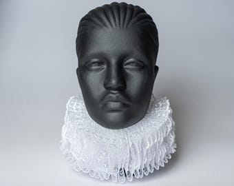 May include: A black bust of a person wearing a white, ruffled Elizabethan collar. The bust has a smooth, matte finish and closed eyes. The collar is made of delicate, layered fabric.