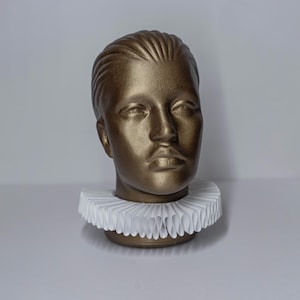 Puede incluir: Un busto de color dorado con un cuello de volantes blanco. El busto es una representación simple y realista de la cabeza y el cuello de una persona.