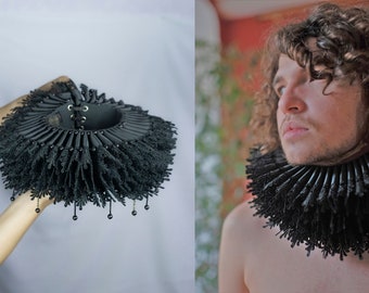 May include: Black ruffled collar with lace trim and beaded accents. The collar has a central opening with a lace-up closure. The image shows the collar being held and worn, showcasing its design and texture.