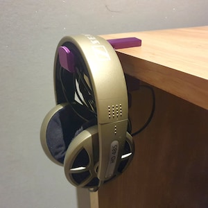 May include: A pair of gold-coloured headphones with black earcups are hanging on a purple desk headphone holder attached to the edge of a wooden desk. The headphones have the text "HD 485" printed on the side.
