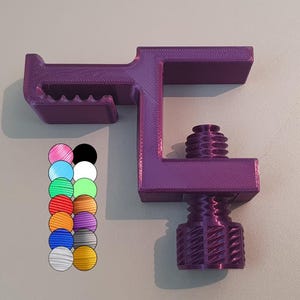 Puede incluir: Una abrazadera de color púrpura impresa en 3D con una perilla roscada. La abrazadera tiene un pequeño labio en un lado y un agujero roscado en el otro.
