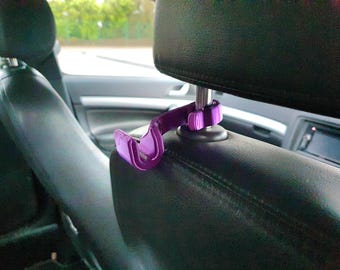 May include: A purple plastic car headrest hook attached to the metal post of a black leather vehicle headrest. The hook features a curved design intended for hanging items and is securely fastened around the headrest support pillar.