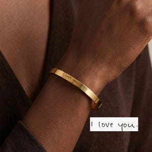 Op de afbeelding: Een goudkleurige metalen armband met de gegraveerde tekst "I love you" in cursief schrift. De armband heeft een gladde, gepolijste afwerking en een eenvoudig open ontwerp. De tekst "I love you." wordt ook weergegeven in een wit kader.
