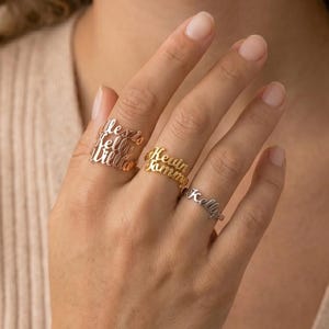 Op de afbeelding: Een hand toont drie gepersonaliseerde naamringen in zilver, goud en roségoud. De ringen zijn voorzien van de namen "Alexis", "Bella", "Lilla", "Kevin", "Tommy" en "Kelly" in een sierlijk lettertype. De ringen worden aan de vingers gedragen.