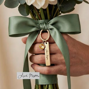 Peut inclure: Un bouquet de dahlias blancs et de feuilles d'eucalyptus noué avec un ruban de satin vert sauge. Une breloque ovale dorée est suspendue au ruban, avec un petit cristal jaune et une plaque rectangulaire gravée du texte Love Mom.