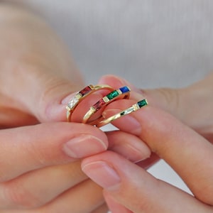 Puede incluir: Cuatro anillos de oro con piedras preciosas de colores. Los anillos están apilados uno encima del otro. Las piedras preciosas son rojas, verdes, azules y transparentes.