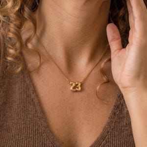 May include: A gold-toned necklace featuring a pendant shaped like the number 23. The pendant has a smooth, rounded, balloon-like appearance and is suspended from a delicate gold-toned chain that rests against the collarbone area.