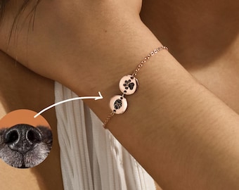 May include: A rose gold chain bracelet with two round charms featuring paw prints. The charms are engraved with a black paw print and a brown nose print.