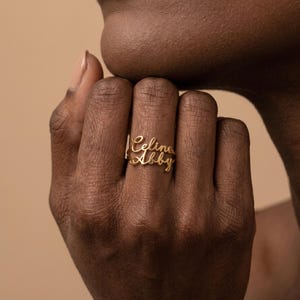 Op de afbeelding: Een goudkleurige ring met twee gestapelde naamplaatjes in cursief schrift. Het bovenste naamplaatje toont de naam Celine in een vloeiend lettertype, terwijl het onderste naamplaatje de naam Abby in een bijpassende cursieve stijl weergeeft.