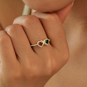 May include: A gold ring with two heart-shaped gemstones, a red and a green one, each encircled by smaller clear stones. The ring sits on a delicate band, displaying a romantic and elegant design.