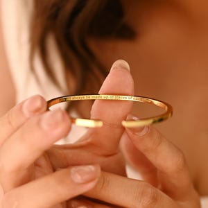 May include: A gold-colored open cuff bracelet. The bracelet has the inscription "will always be made up of pieces of each other" engraved on the outside. The bracelet is held open, revealing the inside.