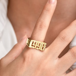 May include: A gold ring featuring the cut-out number "1996" in a distinctive font. The ring has a rectangular shape and is worn on a finger. The background is blurred, highlighting the jewellery.