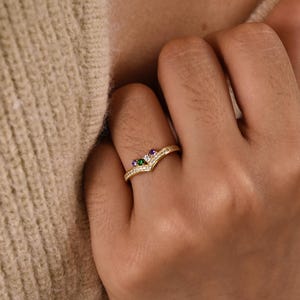 May include: A gold-colored ring with a chevron design. The ring features a small green, purple, and white gemstone cluster at the center, with smaller clear stones along the band. The ring is on a finger.