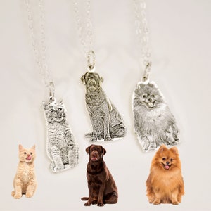 May include: Three silver necklaces with personalised pet portraits. The pendants feature a grey cat, a black dog, and a grey dog. The necklaces are hanging against a white background.
