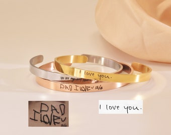 Puede incluir: Tres pulseras de brazalete de oro, plata y oro rosa con mensajes grabados. La pulsera de oro dice "Te amo". La pulsera de plata dice "Eres mi favorito". La pulsera de oro rosa dice "DAD I LOVE U 12/12".
