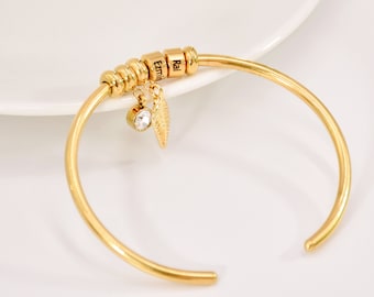 Puede incluir: Pulsera de brazalete de oro con un dije de pluma y un cristal. La pulsera tiene una serie de cuentas con los nombres "Ezm", "Rai" grabados en ellas.