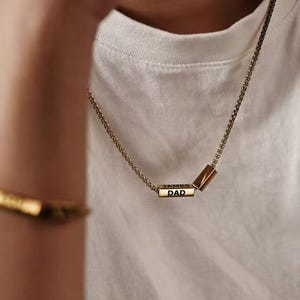 Könnte beinhalten: Goldfarbene Halskette mit rechteckigem Anhänger mit der Aufschrift "DAD". Die Halskette wird mit einem passenden goldenen Armband getragen. Die Person trägt ein weißes Tanktop. Der Anhänger ist etwa 2 cm lang.