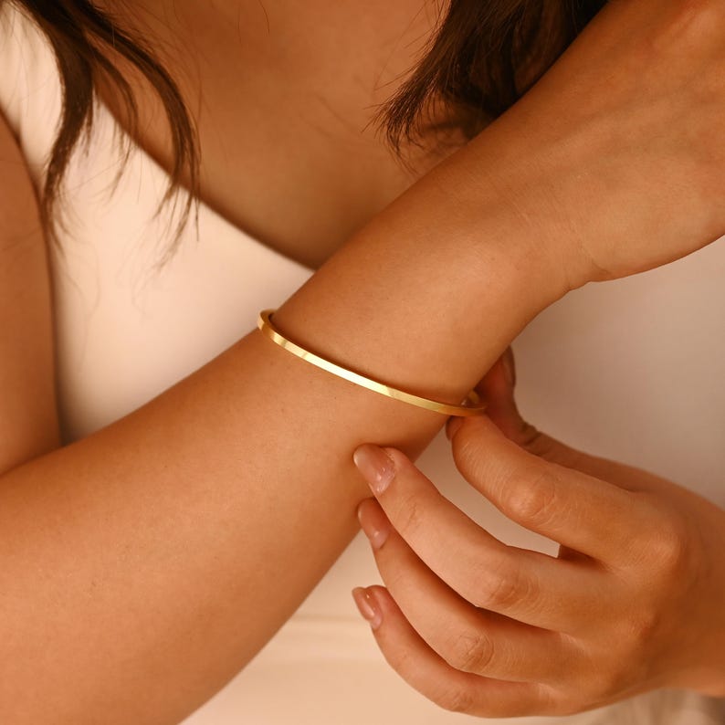 May include: A gold-colored, thin bangle bracelet. The bracelet is a simple, minimalist design, with a smooth, polished surface. The jewelry is being put on a wrist.