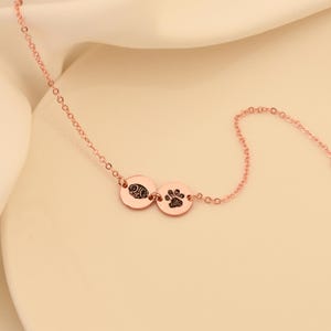 Puede incluir: Un collar de oro rosa con dos colgantes redondos. Un colgante tiene una huella de pata y el otro una huella dactilar.