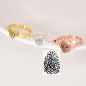 May include: Three rings with fingerprint designs. One ring is gold with a heart-shaped fingerprint, another is silver with a circular fingerprint, and the third is rose gold with an oval fingerprint. A black and white fingerprint image is also shown.