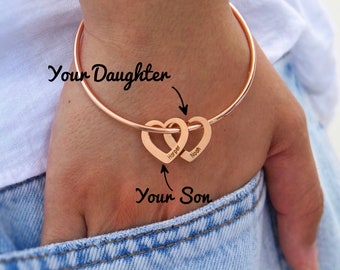 Puede incluir: Pulsera de brazalete de oro rosa con dos dijes de corazón entrelazados. Los corazones están grabados con los nombres "Harper" y "Noah".