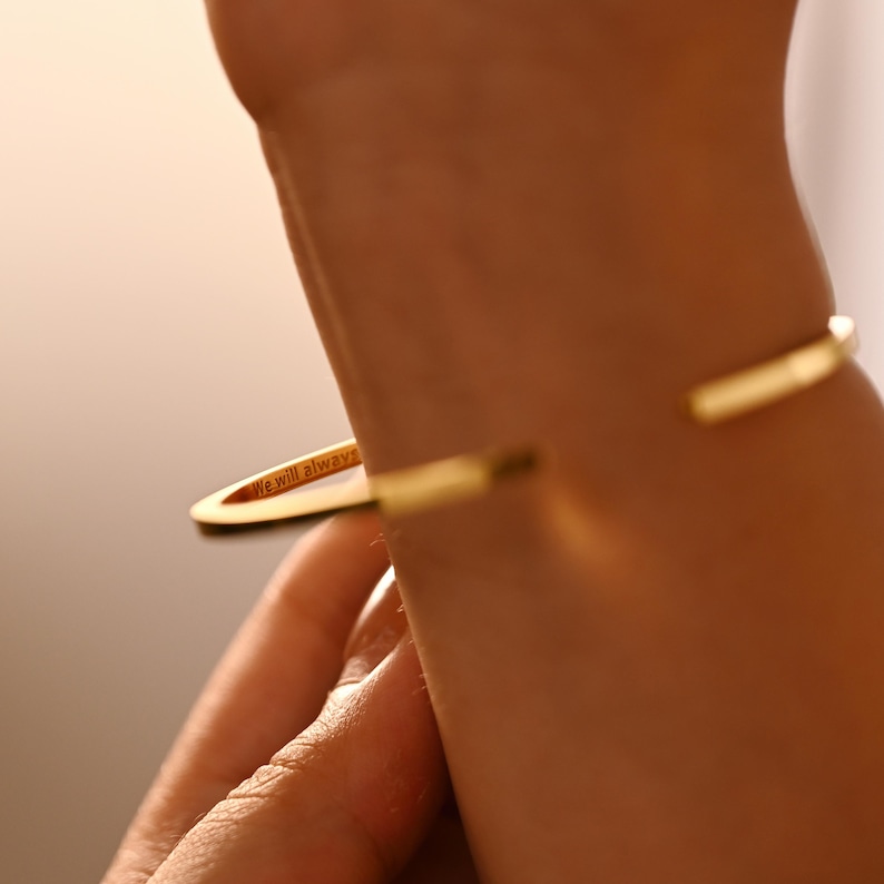 May include: A gold-colored cuff bracelet with the engraved words "We will always" on a flat, oval-shaped accent. The bracelet is open and worn on a wrist.