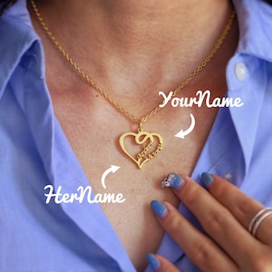 Op de afbeelding: Gouden ketting met een hartvormige hanger met twee namen, "Andrew" en "Victoria", die in elkaar zijn gevlochten. De ketting wordt gedragen door een persoon met blauwe nagellak.