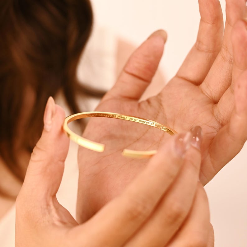 May include: A gold-colored open cuff bracelet. The bracelet has an engraved inscription that reads "We will always be made up of pieces of everything". The bracelet is held in a person's hand.
