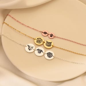 Puede incluir: Tres collares de oro con dijes ovalados que muestran retratos de mascotas y huellas de patas. Los collares est&aacute;n superpuestos sobre una superficie blanca.
