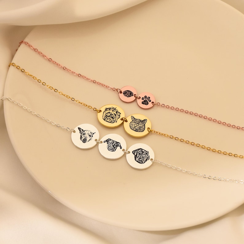 Puede incluir: Tres collares de oro con dijes ovalados que muestran retratos de mascotas y huellas de patas. Los collares est&aacute;n superpuestos sobre una superficie blanca.