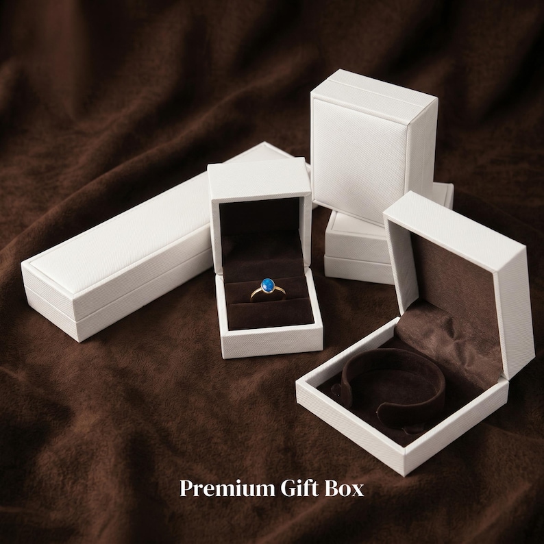 May include: Several white jewelry gift boxes, some open to reveal a ring and a bracelet holder. The ring has a gold band and a blue gemstone. The boxes are on a brown surface. Text on the image reads "Premium Gift Box."
