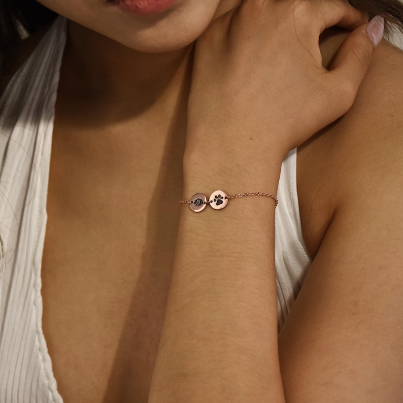Puede incluir: Un brazalete de cadena de oro rosa con dos peque&ntilde;os dijes redondos. Un dije tiene un dise&ntilde;o de huella de pata y el otro dije es liso.