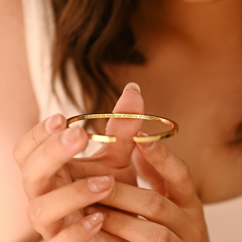May include: A gold-colored open cuff bracelet. The bracelet has the inscription "will always be made up of pieces of each other" engraved on the outside. The bracelet is held open, revealing the inside.