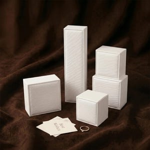 May include: An assortment of white jewelry boxes in various sizes, including a tall rectangular box and several square boxes. A small gold ring and a card with "Best Wishes" are also present. The background is a dark brown, textured fabric.