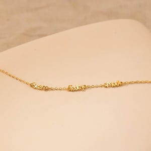 May include: A delicate gold necklace with three cursive name pendants: "Sean", "Fady", and "Madi". The chain and pendants are a warm gold color, creating a simple yet elegant design. The necklace is displayed on a neutral background.